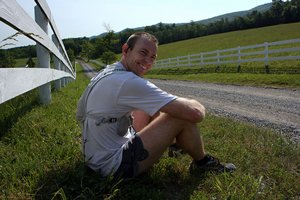 Runner in white shirt, black shorts, sitting in the grass looking at camera smiling.
