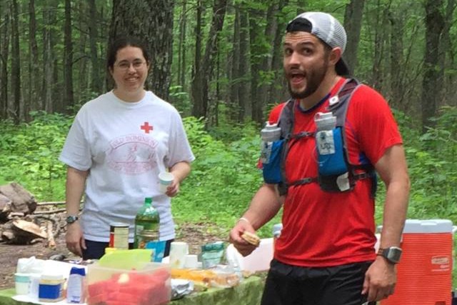Runner in a white hat, red shirt, hydration pack, and black shorts, looks at the camera with a open mouth smile. While 
          a medical team member stands behind a table looking towards the camera.