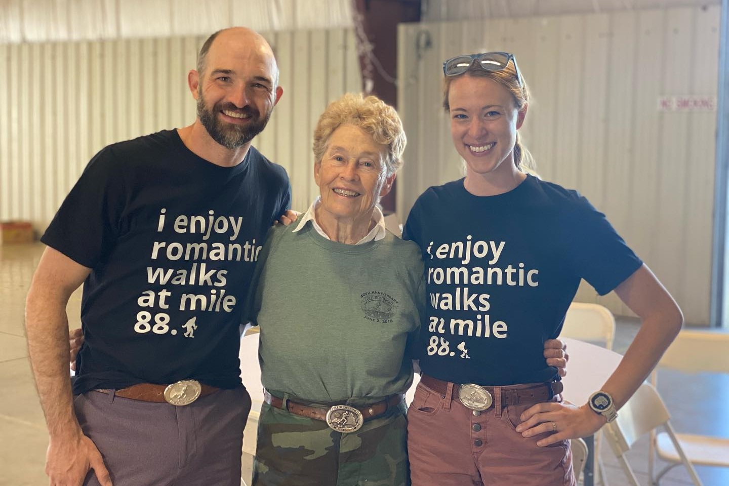 Ben D and Emily L wearing matching shirts that say 'i enjoy romatice walks at mile 88.4' poses with founder, Pat Botts