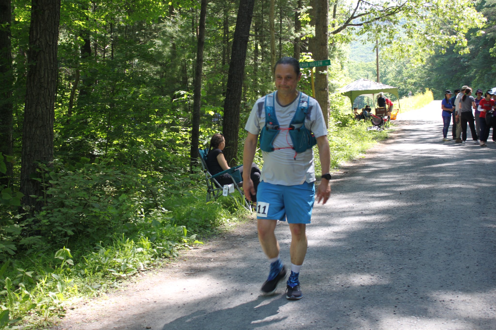 Runner in white shirt, blue hydration pack, and blue shorts, walks into the Aid station