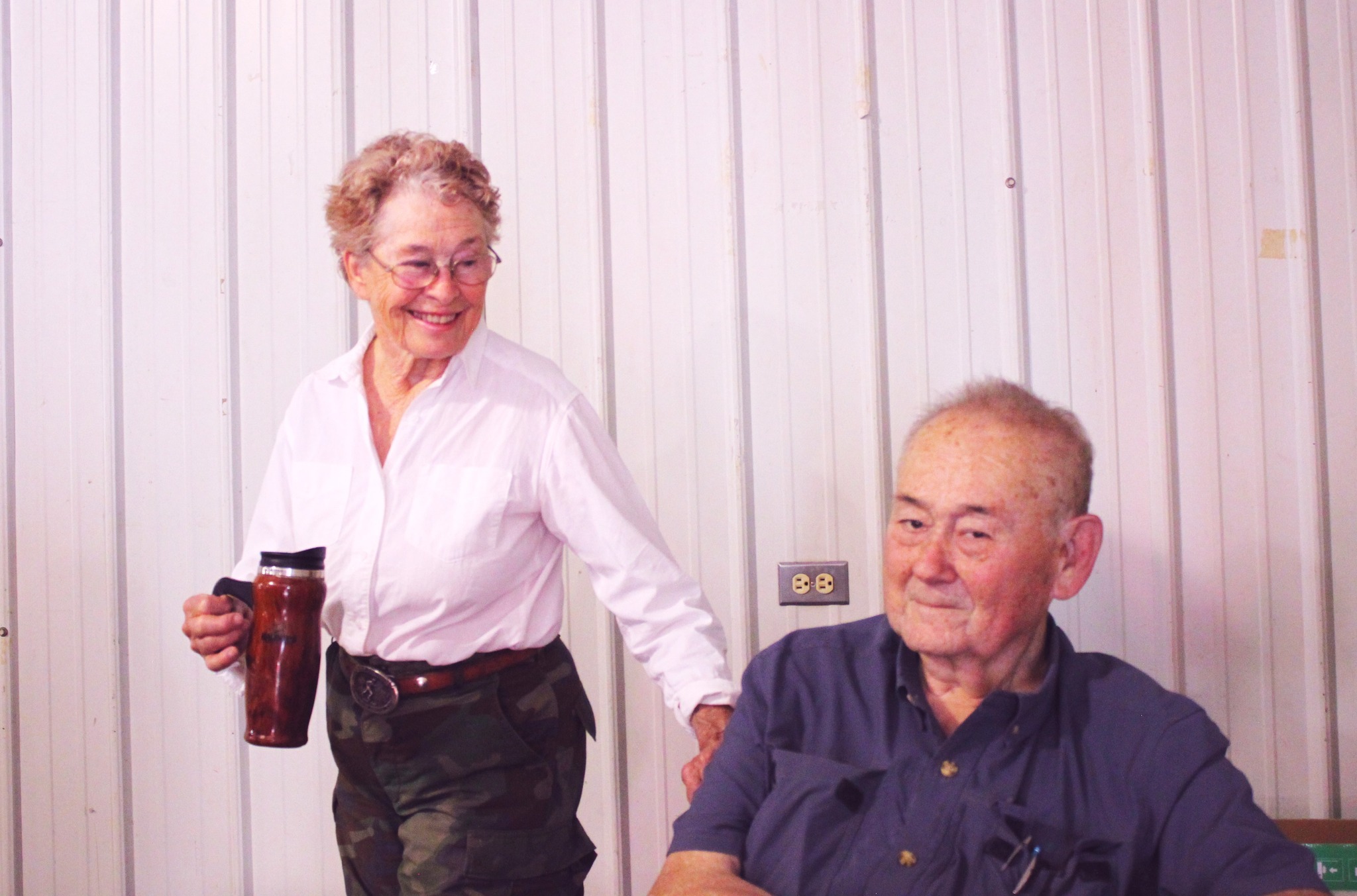 Women on left wears a white blouse holds travel coffee mug while smiling at man on the right. Man on the right wears a blue button down
          shirt and looks at the camera.