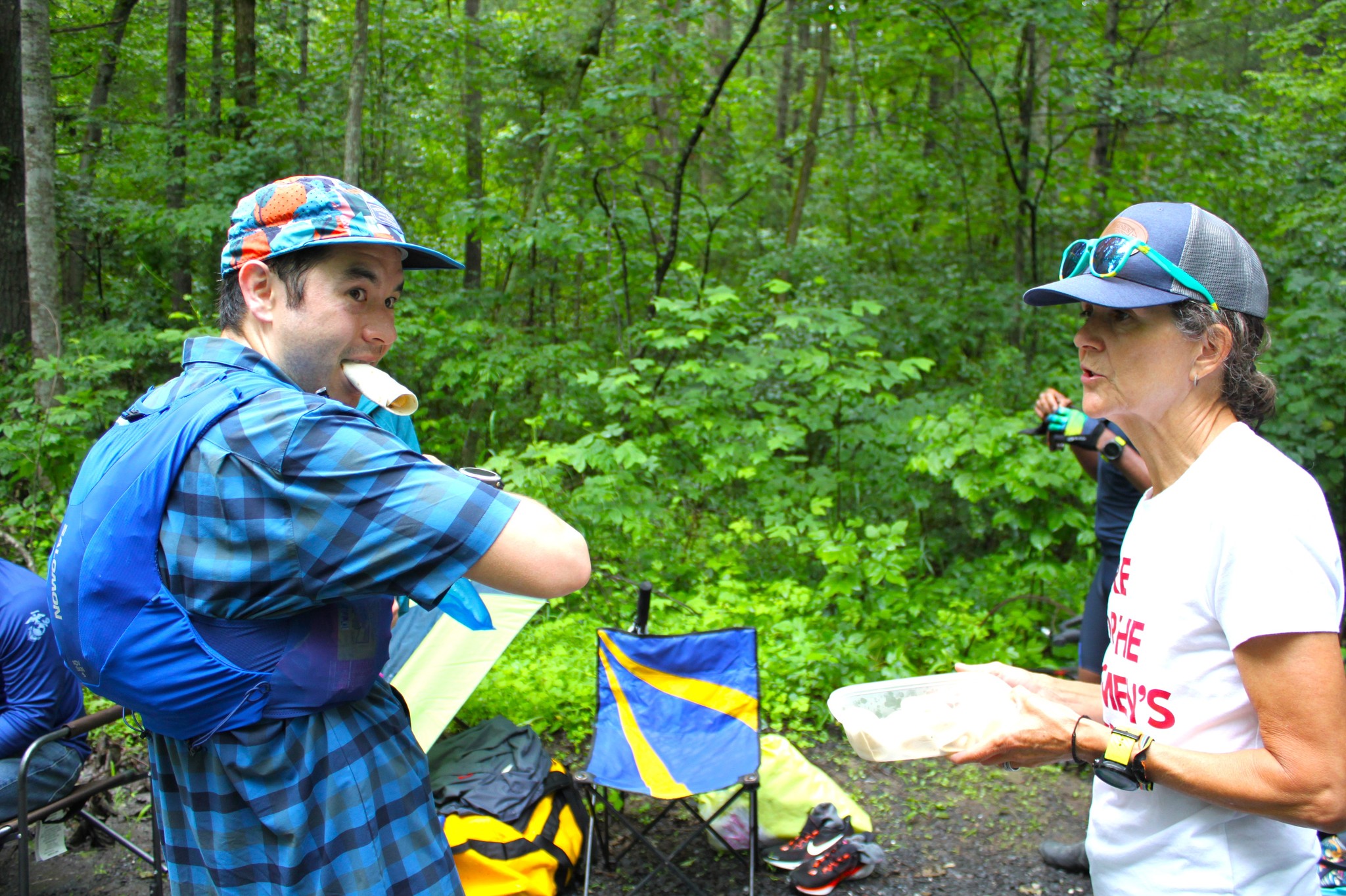 Male runner holds 
            a burrito in his mouth, while grabbing something from his running pack. He looks off to the side, while a crew member
            talks to him.