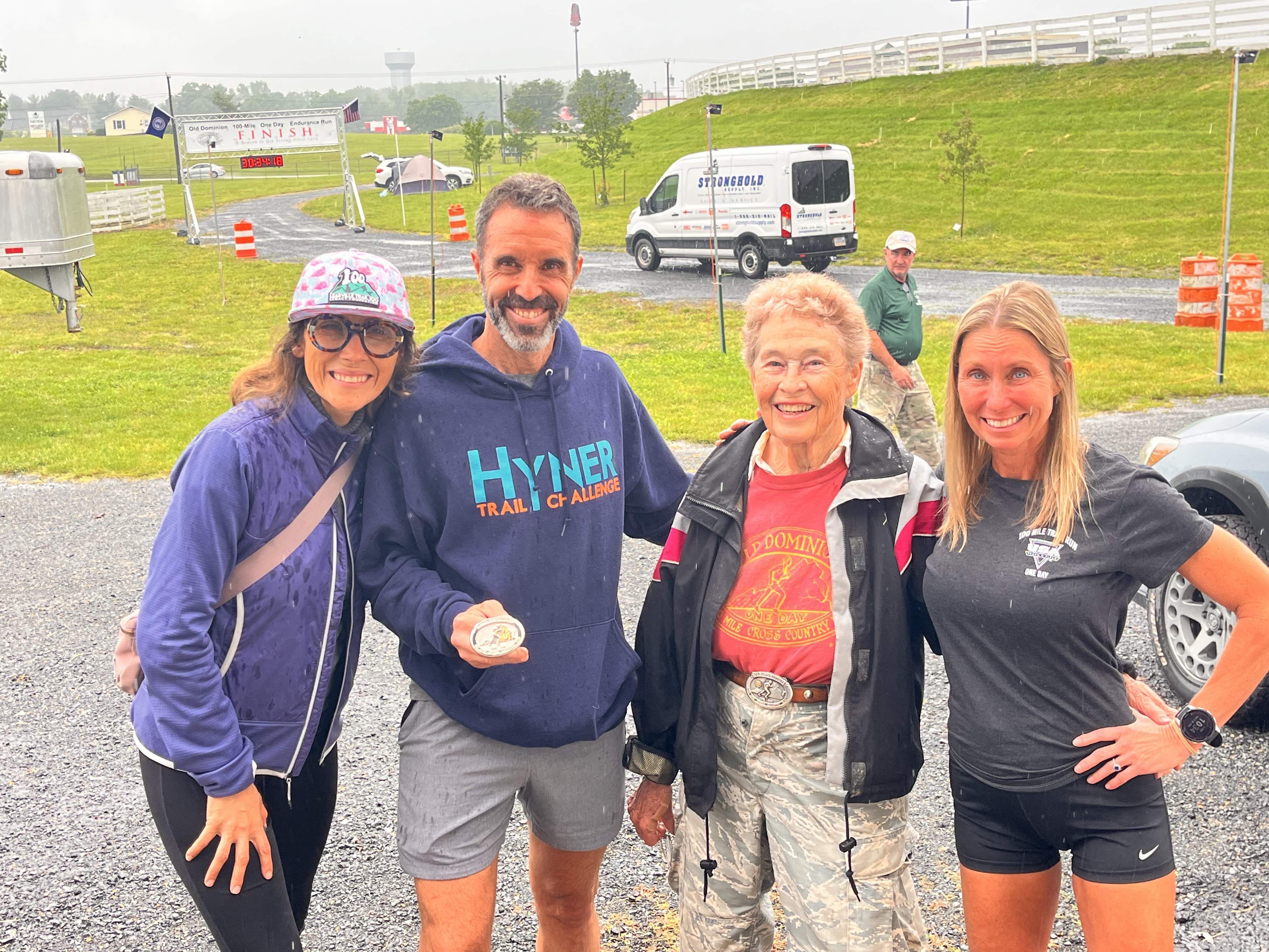 group of athletes stand
        and smile at the camera. Male runner holds a belt buckle.