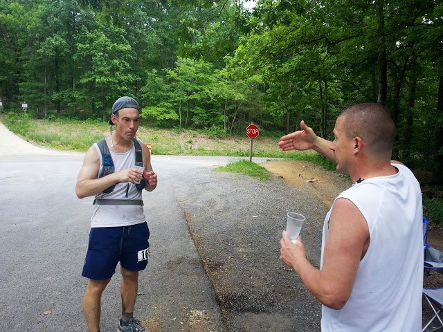 Runner wears a white shirt, blue shorts, hydration vest. He listens closely as another man explains something