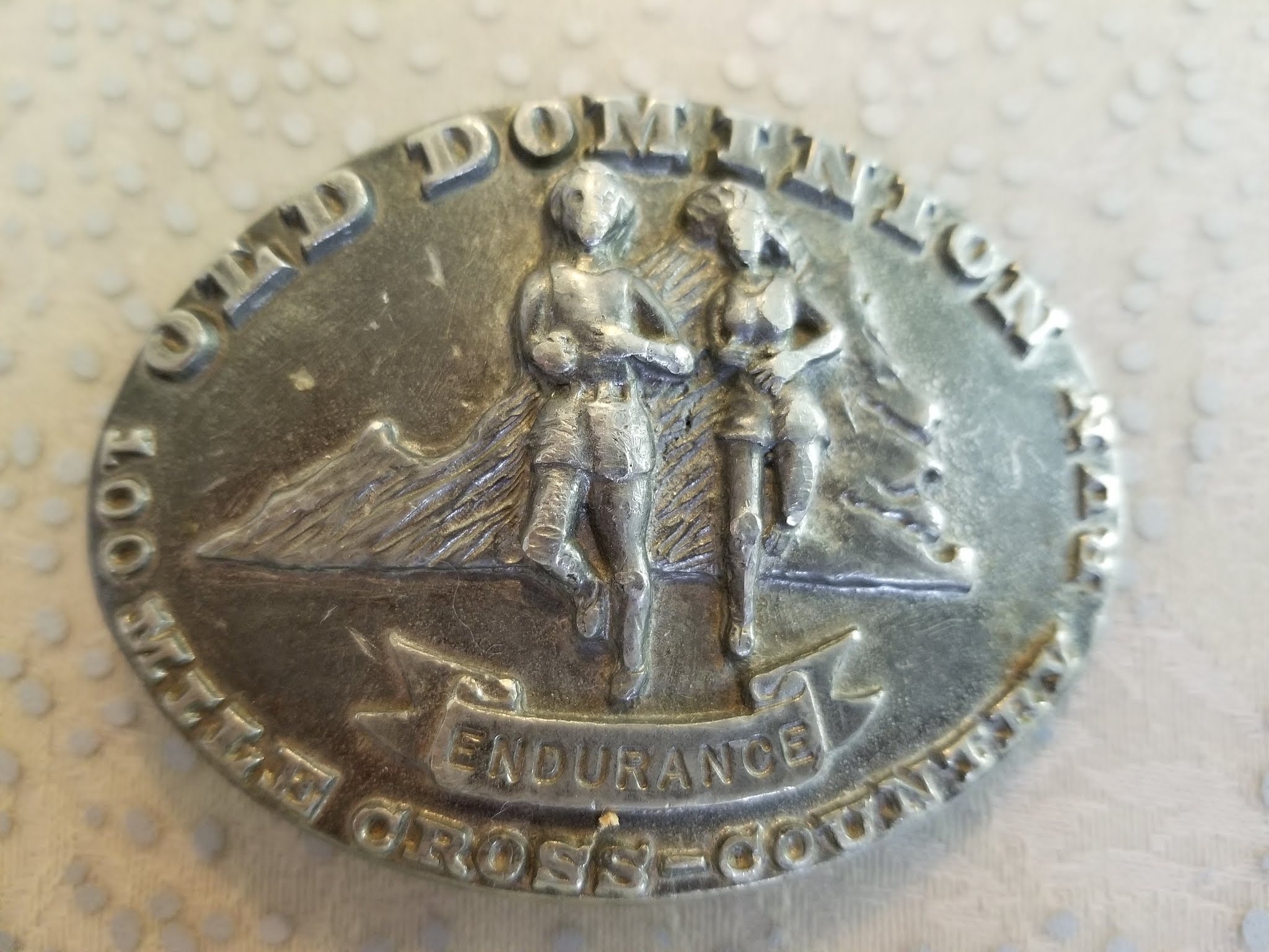 silver belt buckle. Old Dominion across the top, 100 Mile cross-country along the bottom.
             State of virginia in the background. A female and male runner in the foreground
