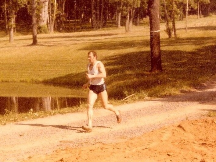 Runner in white shirt, black shorts, running on gravel road with trees in the background