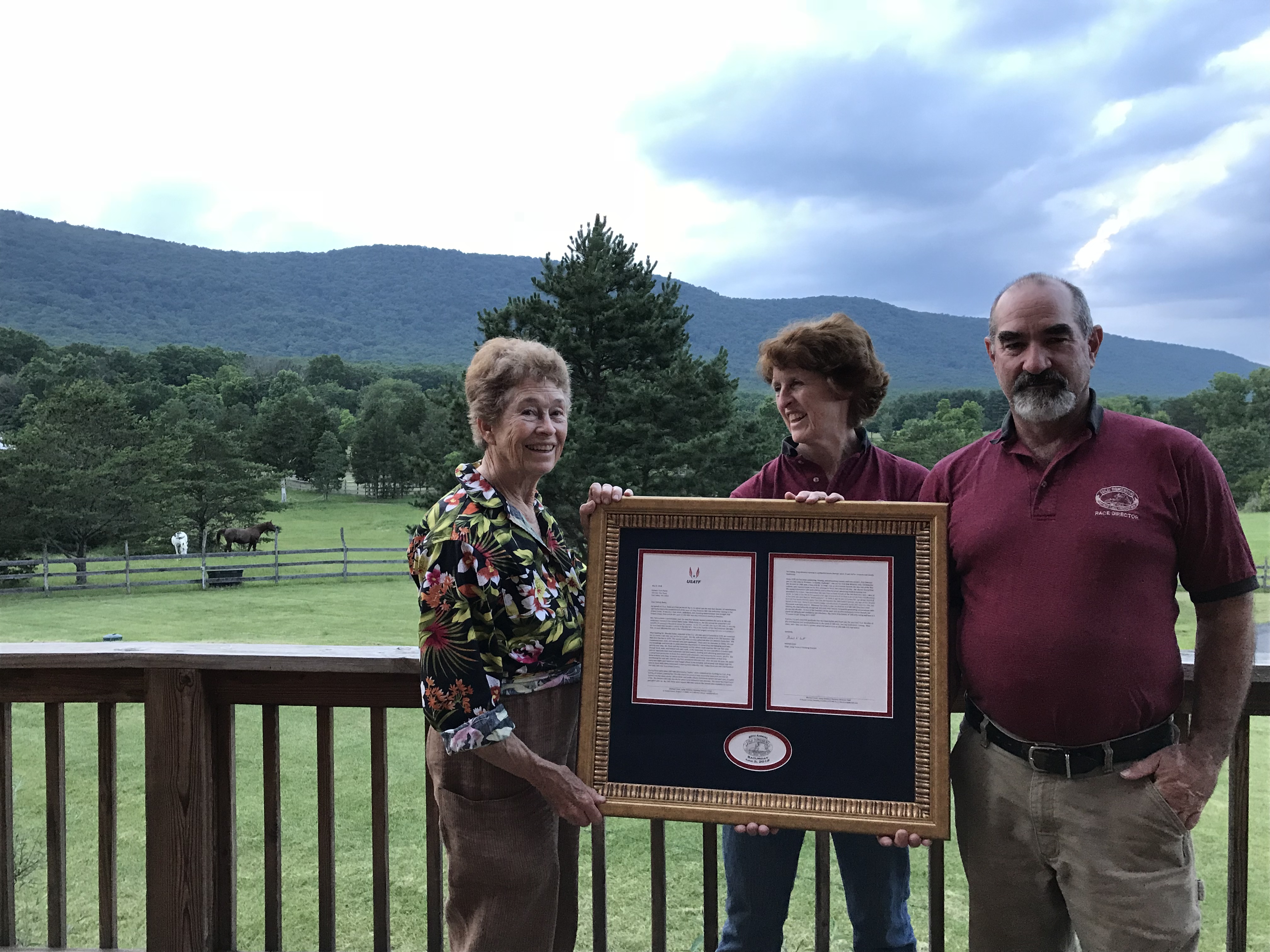 Race Founder Pat Botts holds a frame award on left. Center and right, Current Race Directors support the frame. In the Background 
          is the mountains of the race course.